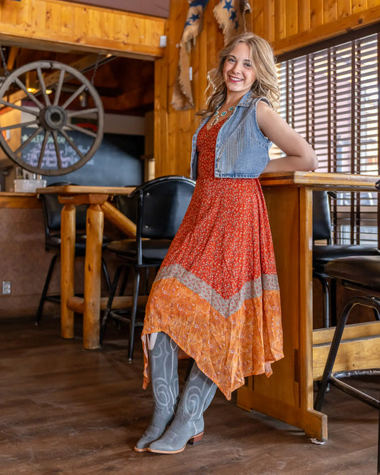 Lacey in a Western saloon wearing a red-orange floral handkerchief dress, light blue cowgirl boots, and a cropped denim vest with fringe — styled for relaxed ranch wear or casual Western occasions.