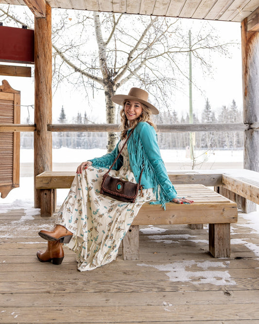 Model in printed maxi dress, turquoise suede fringe jacket, caramel Western boots, and wide-brim hat, sitting on a wood bench.