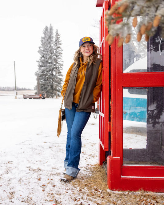Model in Ariat hoodie, Rebar vest, high-rise jeans, and Ariat Southwestern Hilo shoes, standing outside a rustic cabin.