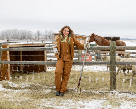 Model in insulated Carhartt bibs, sherpa-lined jacket, and Muck Boots standing in a snowy ranch paddock.