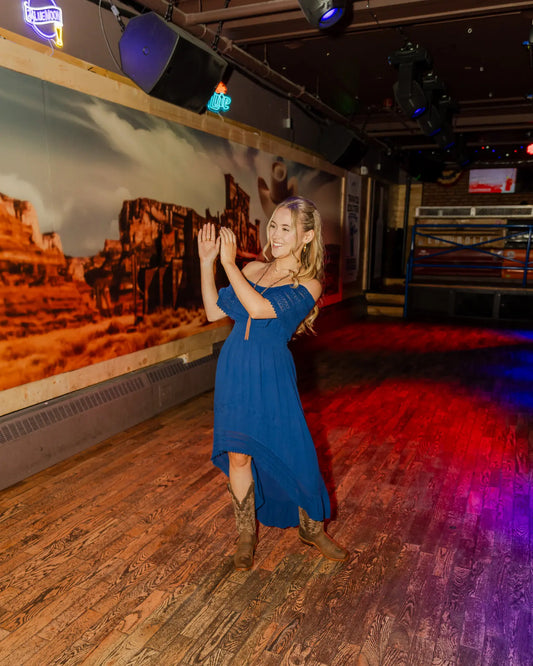 Madeleine Mae smiling and clapping on the dance floor at Daisy’s Saloon, wearing a flowing blue dress with cowboy boots, showcasing western style during the Wei’s Western Wear photoshoot.